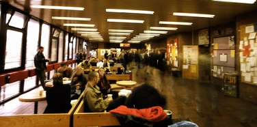 Zeigt den Eingangsbereich vor der Cafeteria mit Blickrichtung zu den Aufzügen. Rechts an der Wand hängen mehrere Anheftfläschen für WG-Gesuche, Praktikumsplätze, Flugblätter etc. Zwischen den Infowänden befindet sich eine Tür, die zur Fachschaft, dem Frauen- und lesbenreferat sowie dem Schwulreferat führt. Links ist eine Fensterfront, die bei schönem Wetter viel Tageslicht spendet. Mittig links stehen Sitzbereiche aus Holz. Einige Studenten sitzen und machen Pause, andere laufen vorüber. Belichtungszeiten bei der Bildaufnahme wurde so eingestellt, dass Bewegungsverwischungen auf dem Bild entstanden sind.