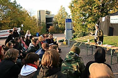 Studierende sitzen draussen im Protestcamp vor einem Tisch, an dem Prof. Schiller referiert