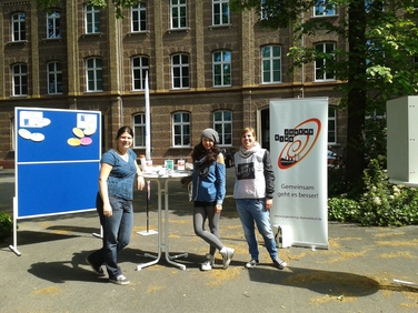 Drei junge StudentInnen stehen auf einem Schulhof. Im Hintergrund sieht man den Stand mit einer Flagge des Jugendrings.